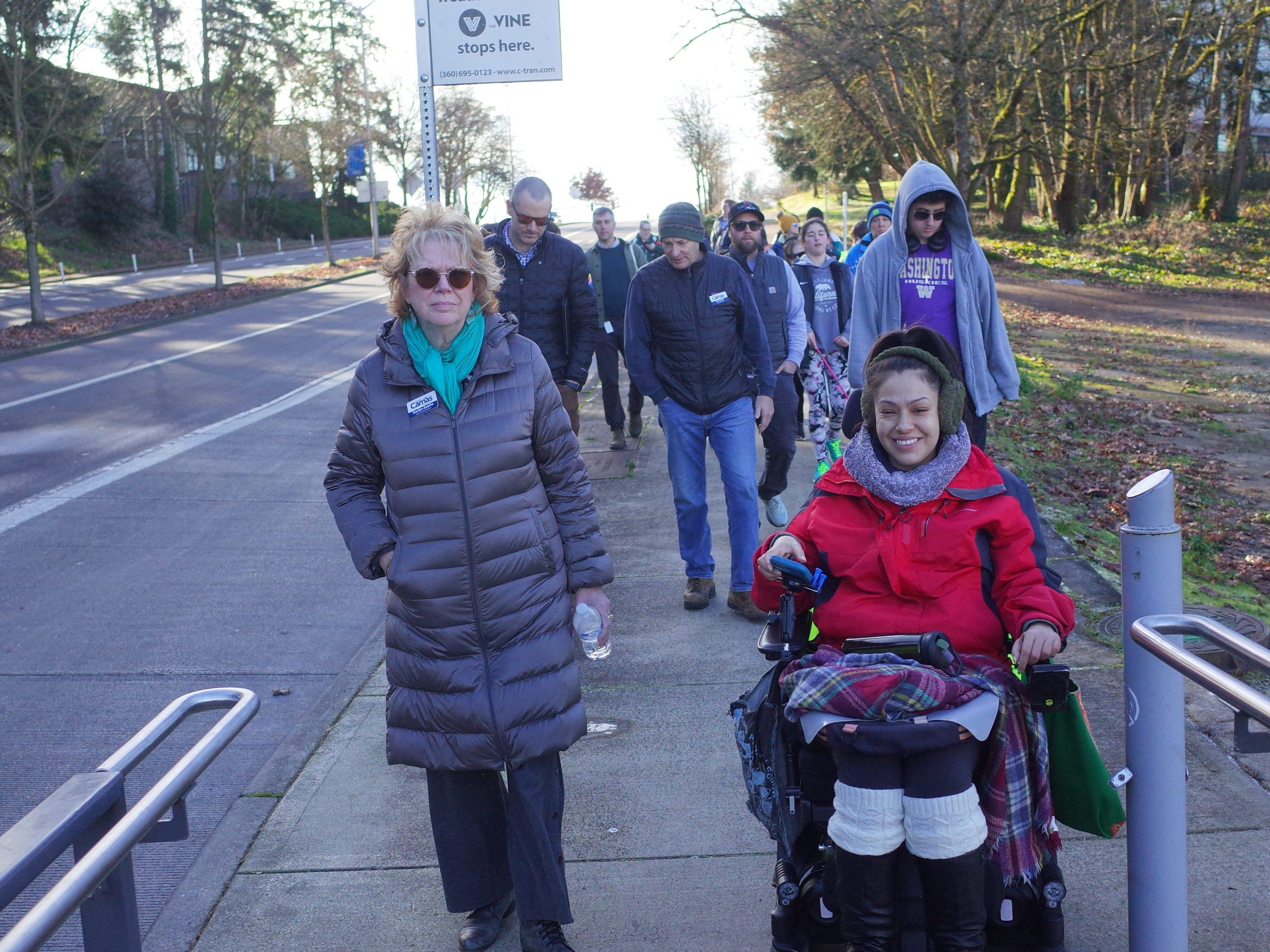 A woman in a gray puffer coat walks along a path next to a road, alongside a child using a power wheelchair in a red jacket. A bike route sign is visible on a post nearby, with evergreen trees in the background.